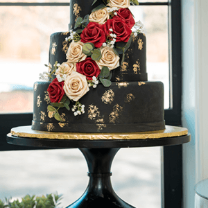 The WD-000C is a three-tiered black cake with gold accents, cascading red and cream roses, greenery, and small white flowers, displayed on a black cake stand by a window.