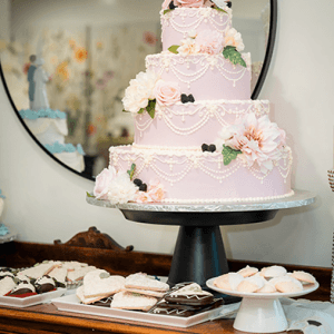The WD-000B three-tiered pink wedding cake with floral and pearl icing is displayed on a black stand, surrounded by cookies and pastries on a wooden table in front of a round mirror.