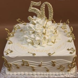 The BD-063 cake features a square white base with gold music notes, white roses, elegant swirls, and a large gold "50" topper. "Happy Birthday Ivano" is piped in gold icing. The cake is set on a patterned base for an elegant finish.