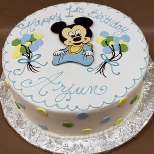 CB-002A: Round birthday cake decorated with baby Mickey Mouse, pastel balloons, blue and green polka dots, and "Happy 1st Birthday Arjun" in blue icing. Presented on a silver foil-covered board.