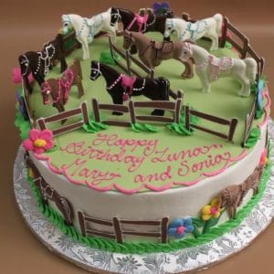 CB-012L: Round birthday cake with green icing, topped with small plastic horses, a brown fence, side flowers, and pink writing that says “Happy Birthday Zina, Mary, and Sonia.”.
