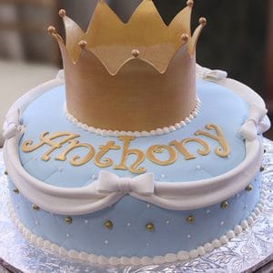 CB-016A is a round cake with light blue fondant, white bows, gold accents, and "Anthony" in gold letters. A large gold fondant crown decorates the top.