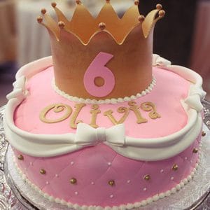 Celebrate a 6th birthday with cake CB-016B: a pink cake featuring gold accents, white bows, the name Olivia, and topped with a gold crown and pink number 6.