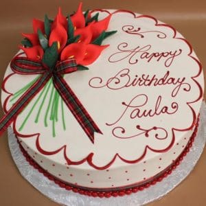 CH-028 is a round white birthday cake featuring red calla lilies, a plaid ribbon bow, red scalloped icing, and "Happy Birthday Paula" in red script. It is presented on a silver board.