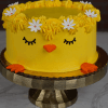 EA-000C is a round yellow cake designed like a chick, featuring closed eyes, an orange beak and feet, with a crown of yellow frosting and white flowers on top, displayed on a cake stand.