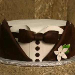The GR-005 is a round cake styled as a tuxedo, featuring brown icing for the jacket, bow tie, and buttons, white icing for the shirt, and a small white flower with green leaves on the side.