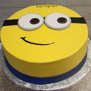The CB-013B is a round cake styled as a Minion with yellow frosting, big white and brown eyes, a black goggles strap, smiling mouth, and a blue band at the base.