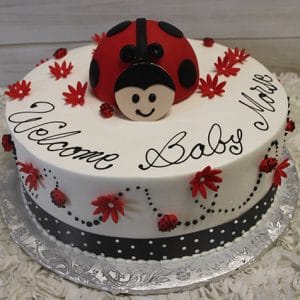CB-012I: Round cake with a large fondant ladybug, red flowers, small ladybugs, and “Welcome Baby Mia” in black icing on white frosting, finished with a black polka dot ribbon around the base.