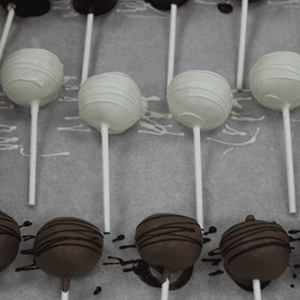 CO-031 Gold Cake Pops are displayed in rows on parchment paper, featuring chocolate and white chocolate pops on sticks, some topped with decorative drizzle.