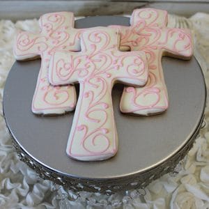 Three CE-005a religious cross-shaped cookies with white icing and pink swirls sit on a round silver stand with a lacy edge, displayed on ruffled white fabric.