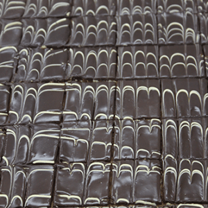Neatly arranged CO-004 Boston Cream 1X1 Bites feature glossy chocolate slices, each topped with a wavy white icing pattern. The uniform bites are displayed in a grid, creating an appealing and elegant dessert presentation.
