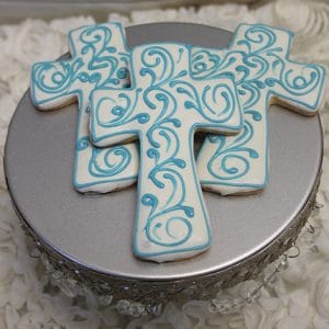 Three CE-005b religious cross-shaped cookies with white icing and light blue swirl designs are displayed on a silver cake stand, accented by white decorative elements.