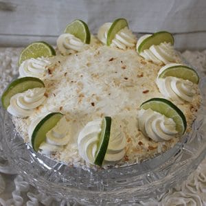 A Key Lime Pie topped with whipped cream swirls, lime slices, and toasted coconut flakes is displayed on a clear glass cake stand.