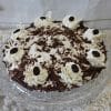 The Pie-Chocolate Cream is a round chocolate pie topped with whipped cream swirls, chocolate shavings, and dark chocolate pieces, displayed on a glass cake stand against a white background.