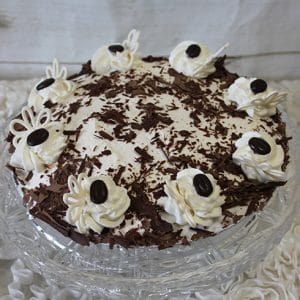 The Pie-Chocolate Cream is a round chocolate pie topped with whipped cream swirls, chocolate shavings, and dark chocolate pieces, displayed on a glass cake stand against a white background.