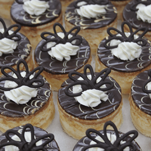 Rows of CO-022 3" Boston Cream pastries topped with glossy chocolate glaze, white cream swirls, and intricate dark chocolate loop decorations.