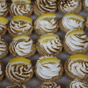 CO-065 Bite Size Mini Lemon Meringue Pies are arranged neatly on a tray, each topped with swirled, torched meringue and a small lemon wedge.