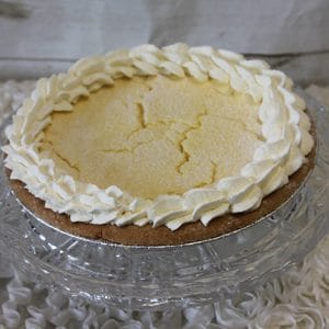 Pie-Ricotta, with a cracked yellow filling and a decorative whipped cream border, sits on a glass cake stand against a light textured background.