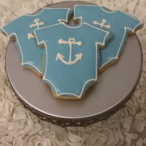 Three CE-004h baby shower cookies, blue and shaped like baby onesies with white anchor designs, are displayed on a round silver stand atop a textured white surface.