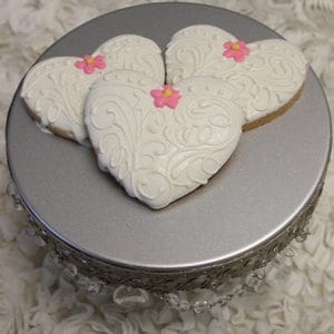 Three CE-003c bridal heart-shaped cookies with white ornate icing and pink flowers are displayed on a silver cake stand with decorative trim, set against a textured white background.