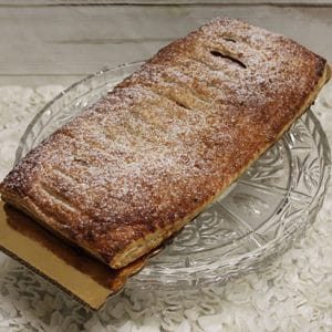 A large rectangular pastry with a golden, flaky crust dusted with powdered sugar sits on a clear glass cake stand. The pastry has a few slits on top and is partially resting on a piece of cardboard.