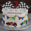 The CB-027D is a round cake featuring colorful race cars, checkered flags, pennant banners, a “Two Fast, Two Curious” topper, and the name Anthony in red icing.