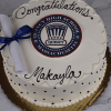 A round white cake (GC-001C Quincy High School) features "Congratulations Makayla" in black icing, a Quincy High emblem at the center, and a rolled diploma with a blue ribbon on top. Displayed on a gold board.