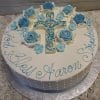RC-053: Round white cake featuring blue and white flowers, a blue ornate cross topper, and "God Bless Aaron Stephen" in blue icing around the edge. Presented on a silver board.