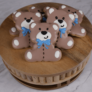 The CE-004d baby shower features bear-shaped cookies with brown icing, white paws, and blue bows arranged on a round wooden board over a light marble surface.