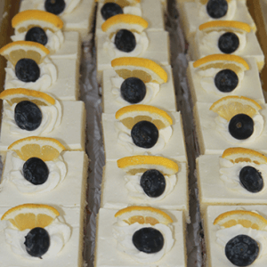 Rows of CO-074 Lemon Blueberry Squares, each topped with whipped cream, a blueberry, and a lemon wedge, arranged neatly on a tray.