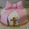 RC-058 is a round white cake with a quilted pattern, gold dots, a gold cross, and a large pink fondant bow. Pink fondant ribbons read "God Bless Ashley." The cake is presented on a gold board.