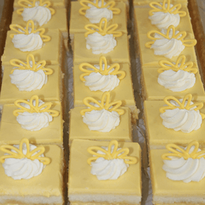 Rows of CO-073 Mango Squares—square yellow cakes topped with a swirl of white whipped cream and yellow icing—are arranged neatly on a tray.