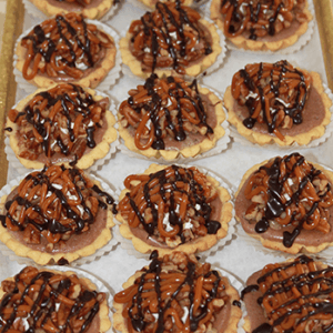 A tray of CO-076 Turtle Cheesecake mini tarts with chocolate and nuts filling, topped with caramel and chocolate drizzle, each served in a white paper cup.