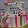 The TC-130 graduation cake features pink and white flowers, gold and pink stripes, a fondant diploma and cap, and “Shekinah Class of 2024” written in black icing.