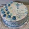 RC-017 is a round white cake adorned with blue and white roses, a blue cross, and the message “God Bless Thomas Michael” in blue writing. Small blue flowers and elegant piped designs add charming accents.