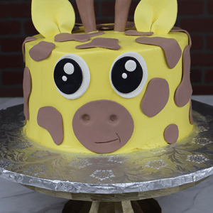 The CB-012E is a round yellow cake designed as a cartoon giraffe face with big eyes, brown spots, fondant ears, and horns, shown on a metallic cake stand against a brick wall background.