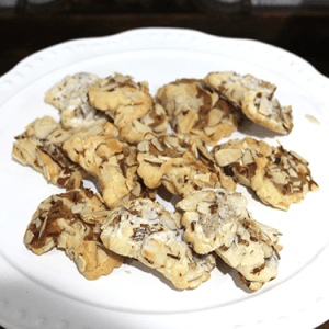 A white plate with several CO-048 Almond Bow tie cookies, topped with sliced almonds, on a dark wooden surface.