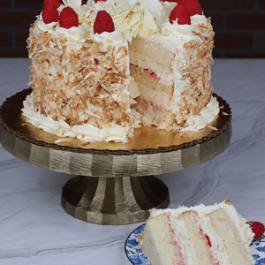 The BD-018Q Raspberry Coconut features three layers of white cake with toasted coconut sides, topped with white chocolate curls and raspberries, displayed on a gold stand. A slice is served on a floral plate in front.