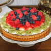 The GD-023 Fruit Tart is displayed on a white cake stand, topped with kiwi, strawberries, raspberries, and blueberries arranged in colorful circles over a smooth creamy filling.