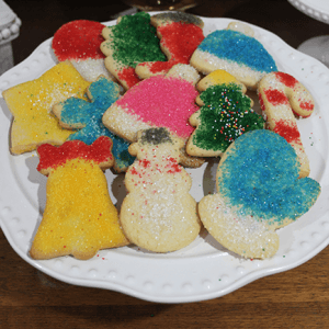 A white plate holds CE-100a Assorted Sugar Christmas Cookies shaped like bells, snowmen, trees, angels, and candy canes, each decorated with colorful sugar sprinkles.