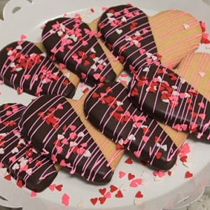 CE-020d Valentine's features heart-shaped cookies half-dipped in chocolate, topped with pink and white drizzle plus heart sprinkles, arranged on a white scalloped plate.
