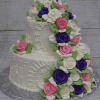 The TC-002B features a two-tiered white cake adorned with detailed piping and fondant roses in purple, pink, and white with green leaves, presented on a silver cake board against a rustic wood background.