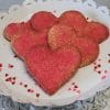 CE-020c Valentine's heart-shaped cookies with red-pink sugar sprinkles are arranged on a white scalloped tray, decorated with small heart confetti and set on a textured white surface.