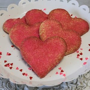 CE-020c Valentine's heart-shaped cookies with red-pink sugar sprinkles are arranged on a white scalloped tray, decorated with small heart confetti and set on a textured white surface.