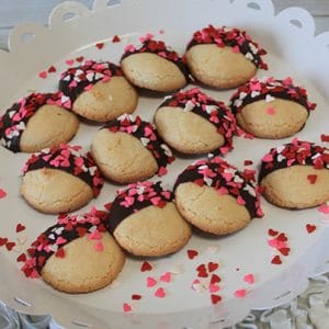 The CE-020g Valentine's features cookies half-dipped in chocolate and topped with red, pink, and white heart-shaped sprinkles on a white scalloped tray, with extra sprinkles scattered around.
