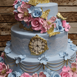 The WD-037 is a two-tier blue cake adorned with pink and blue flowers, gold butterflies, lace-like bands, gold chains, a gold crown topper, and a clock face on the front, displayed against a rustic wooden background.