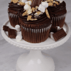 Three CC-000C S'mores cupcakes topped with chocolate frosting, mini marshmallows, chocolate pieces, and graham cracker crumbs are arranged on a white cake stand, with extra chocolate and marshmallows scattered around the base.
