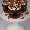 Three CC-000H Chocolate caramel pretzel cupcakes feature white frosting, topped with pretzels and chocolate drizzle, arranged on a white cake stand with scattered pretzel pieces.