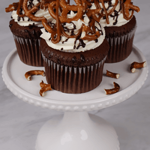 Three CC-000H Chocolate caramel pretzel cupcakes feature white frosting, topped with pretzels and chocolate drizzle, arranged on a white cake stand with scattered pretzel pieces.