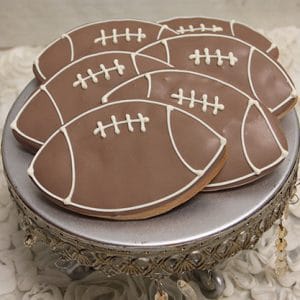 CE-002a sports: Football-shaped sugar cookies with brown and white icing, displayed on a decorative silver cake stand with crystal accents against a textured white background.